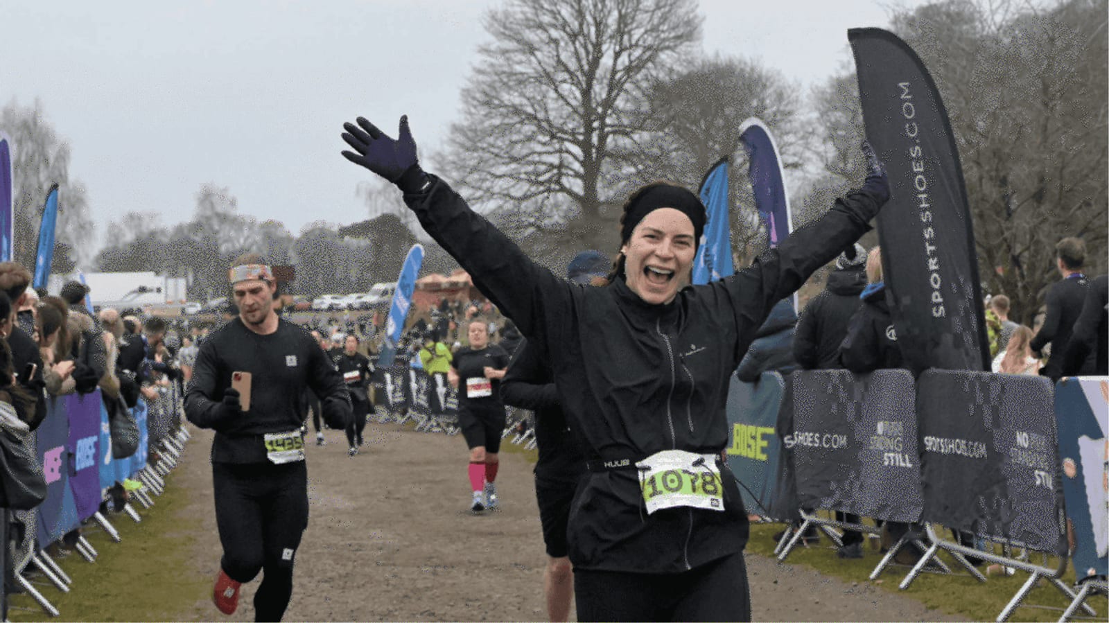 A woman wearing a black outfit and green race bib 1078 smiles with arms raised as she crosses a race finish line, with other runners and cheering spectators behind barriers on a cloudy day.