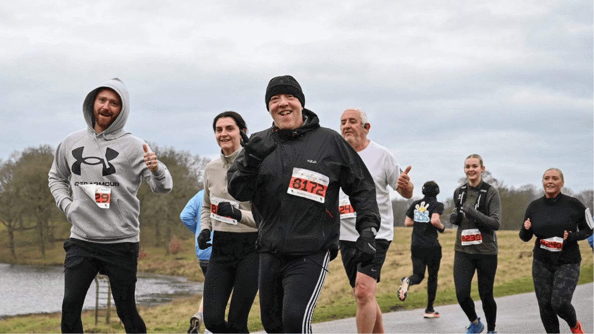 A group of smiling runners wearing race bibs jogs outdoors on a cloudy day, some giving thumbs up, with a river and trees in the background.