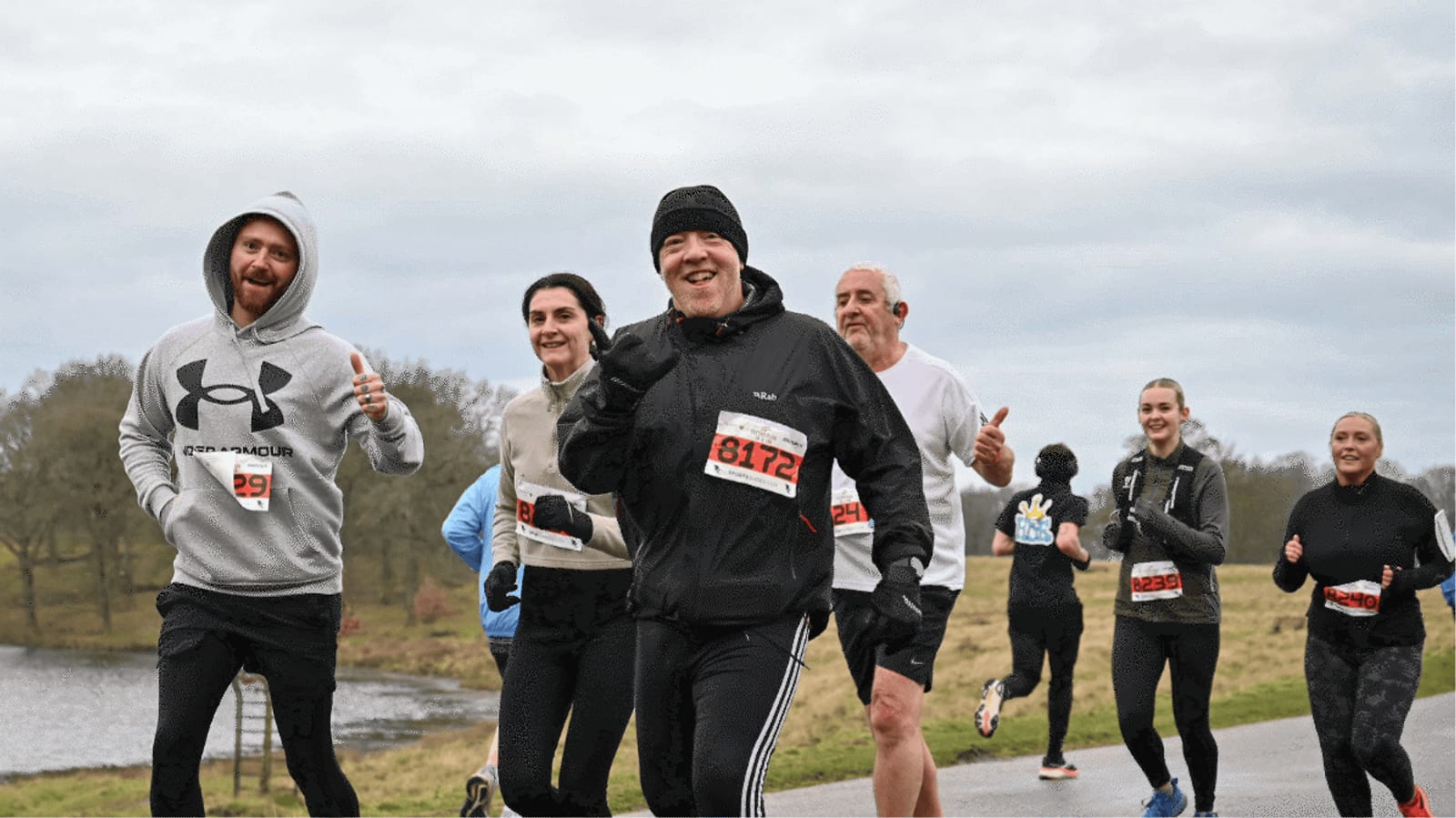 A group of smiling runners wearing race bibs jogs outdoors on a cloudy day, some giving thumbs up, with a river and trees in the background.