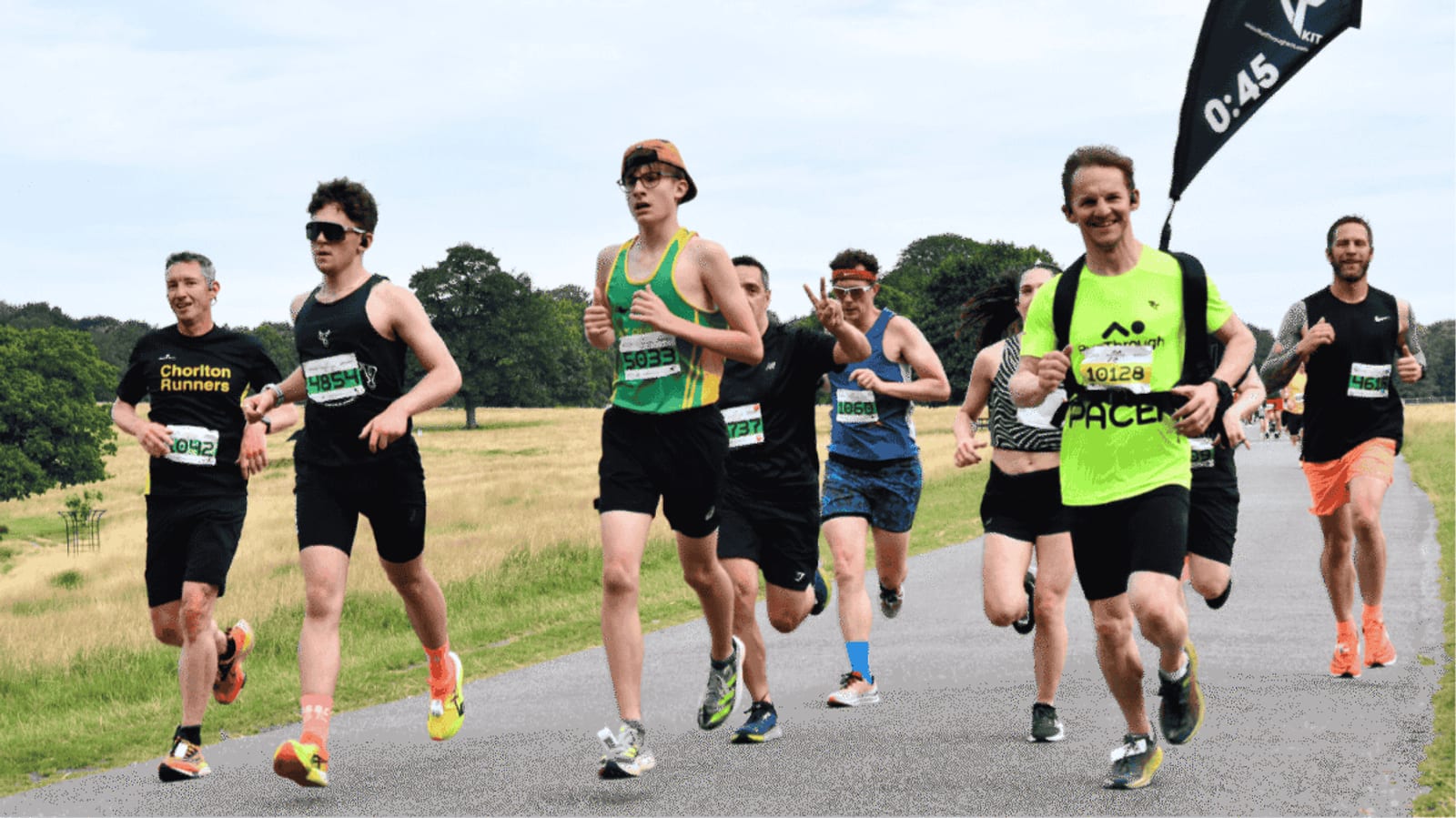 A group of runners, wearing athletic gear and numbered bibs, participate in an outdoor race on a paved path with grassy fields and trees in the background. A black "0:45" flag is visible on the right.