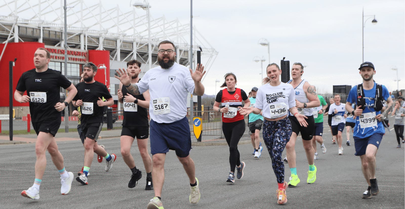 A group of runners participate in a road race near a stadium. Some wave at the camera, while others focus ahead. They wear numbered bibs and athletic clothing on a cloudy day.