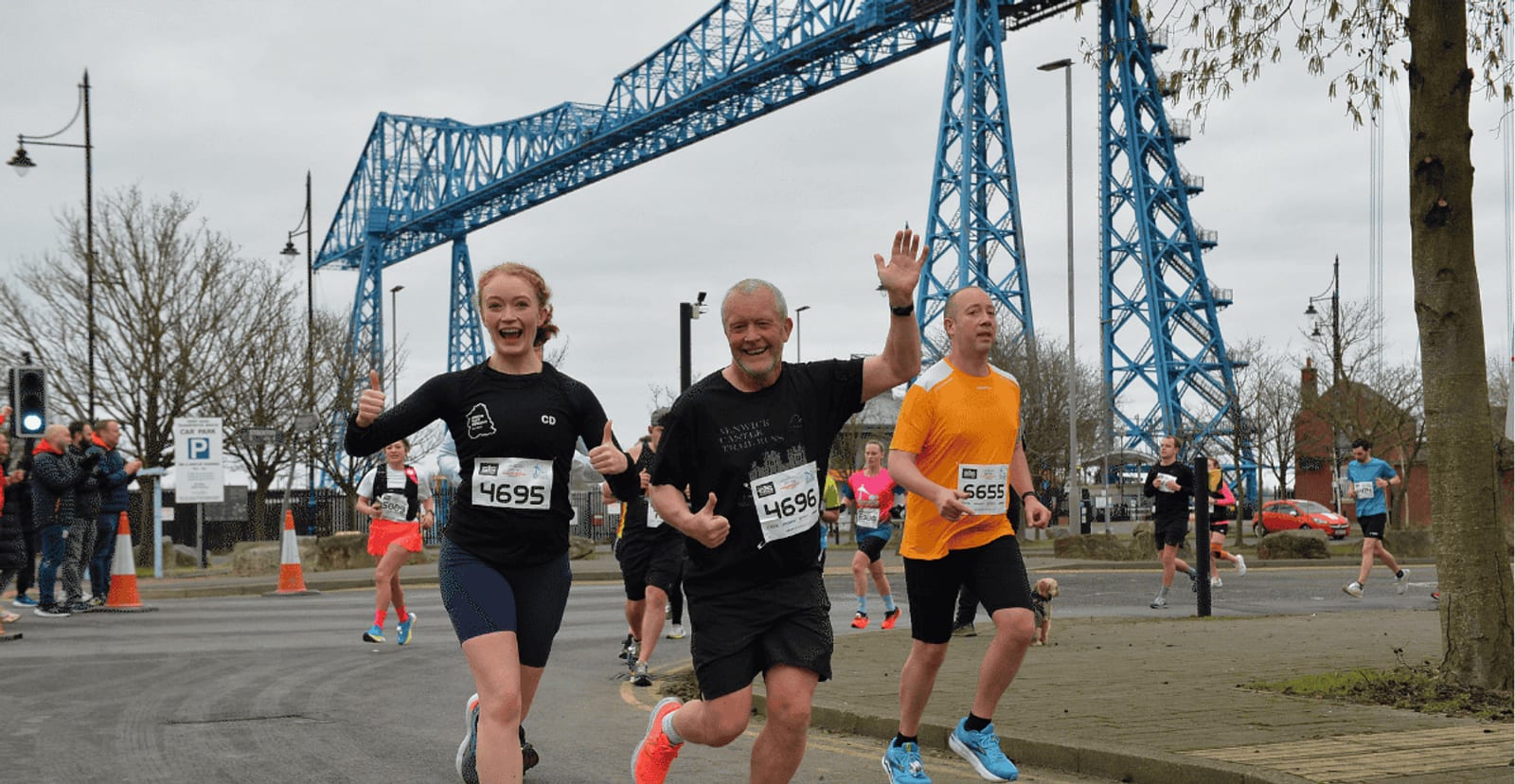 Three runners, two smiling and waving, participate in a road race near a large blue steel bridge on an overcast day. Spectators and other runners are visible in the background.