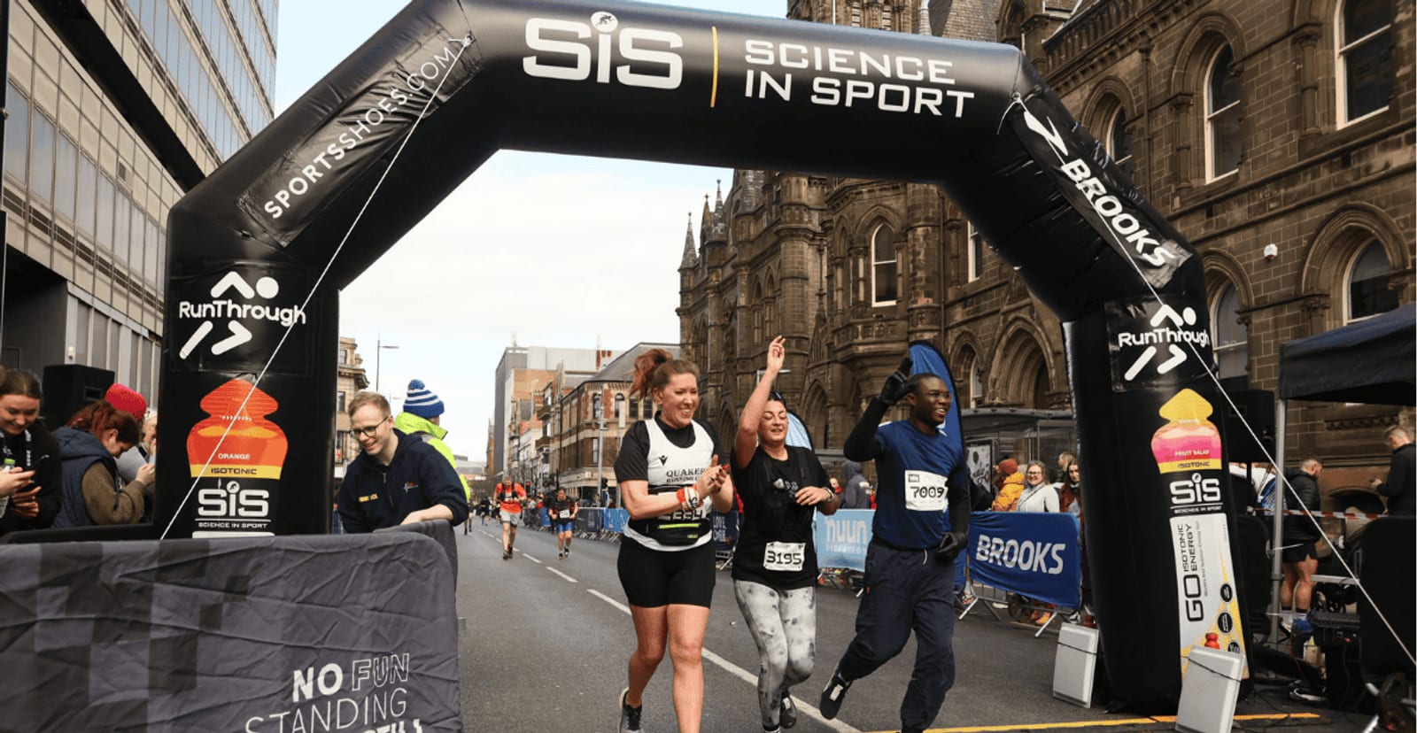 Three smiling runners cross a finish line under a black "Science in Sport" archway on a city street, surrounded by cheering spectators and historic buildings.