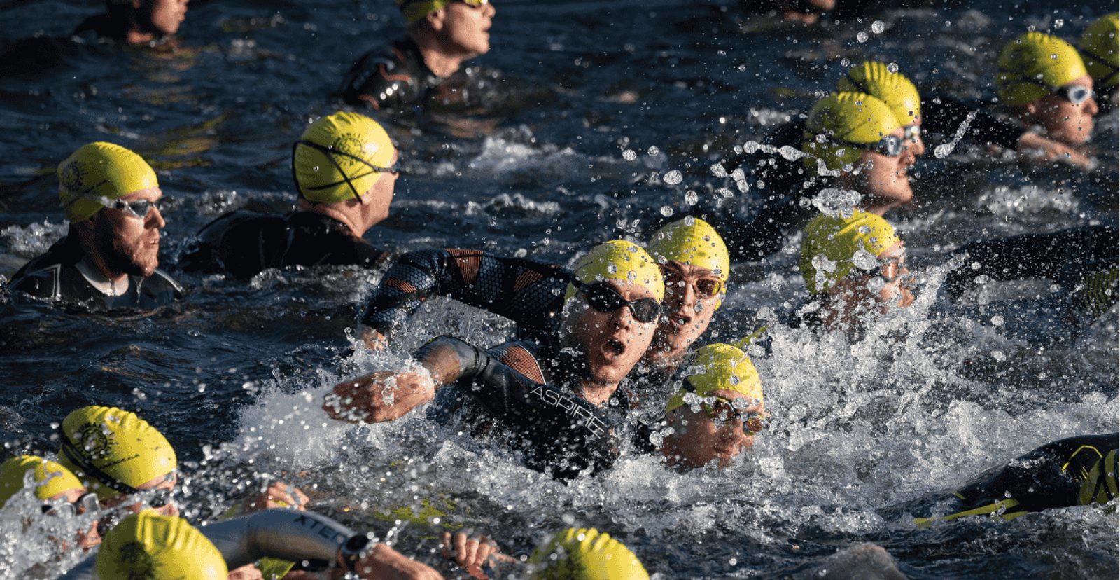 A group of triathletes wearing yellow swim caps and black wetsuits swim close together in open water, creating splashes as they compete in a race.