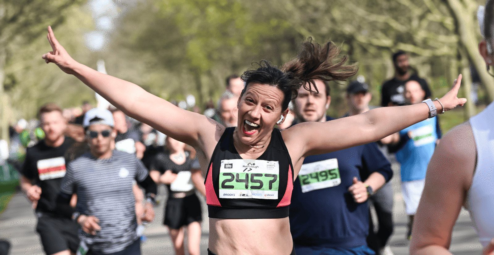 A smiling runner with arms outstretched celebrates while running a race outdoors, surrounded by other participants and trees in the background.