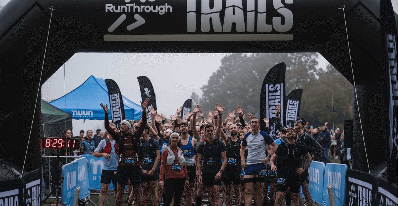 A large group of runners gather at the starting line under a black inflatable archway for a trail race, some raising their hands. Blue race banners and a digital timer are visible. Spectators stand in the background.