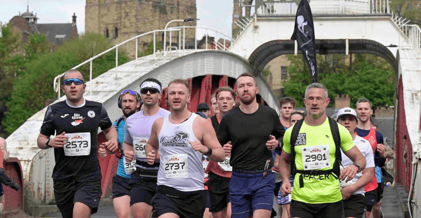 A group of runners, wearing race numbers and athletic gear, run together across a bridge during a race. The background shows a white bridge structure and trees.