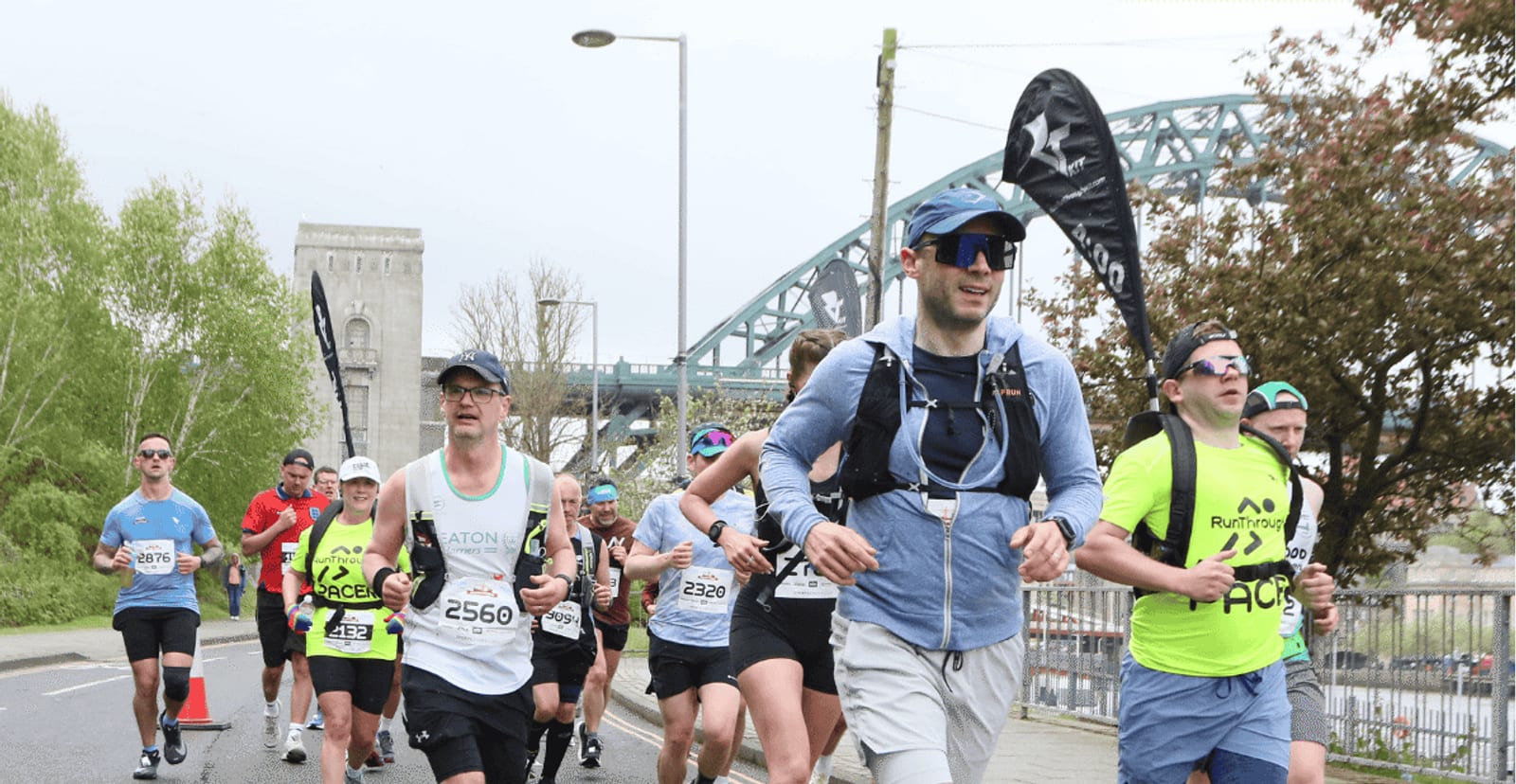 A group of runners in athletic gear participate in a road race on a city street, with a bridge and trees in the background. Some wear sunglasses and hats; race numbers are visible on their shirts.