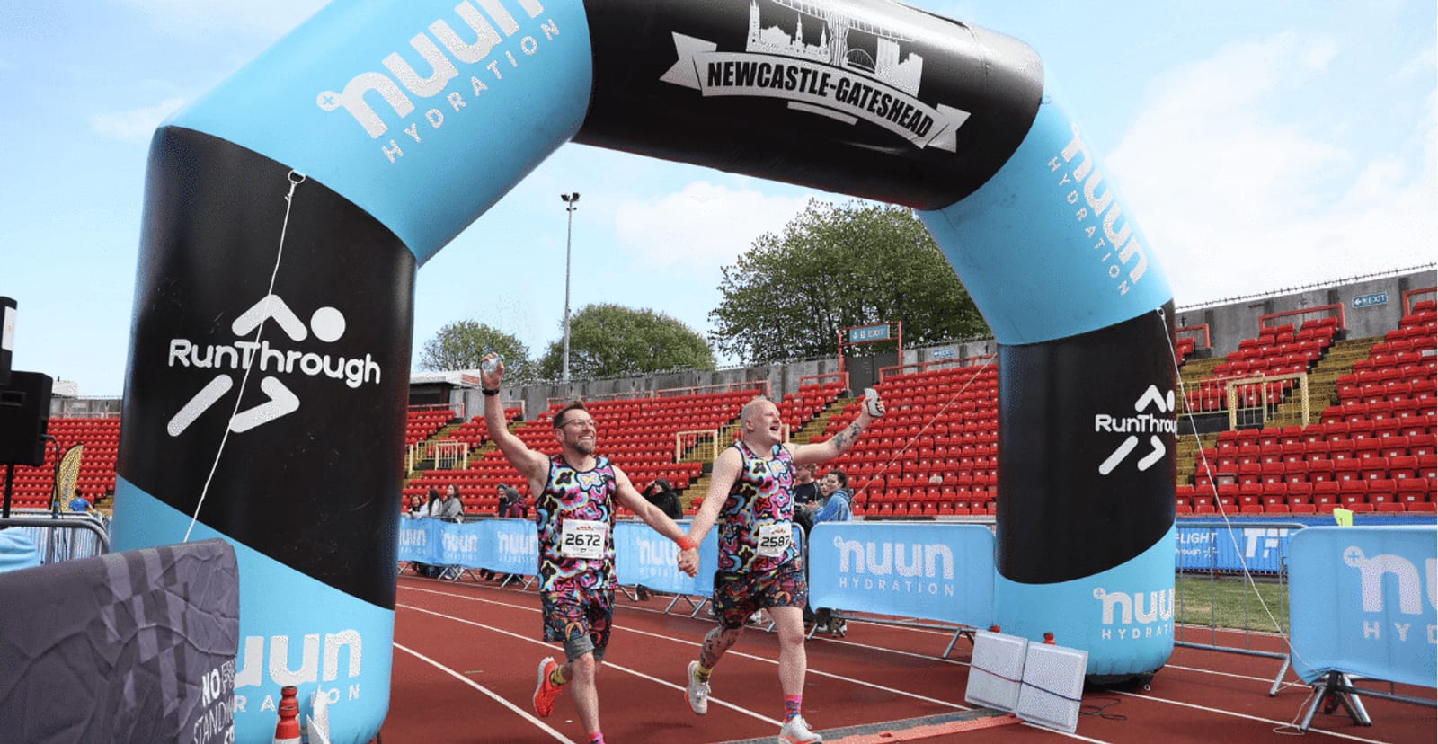 Two runners in colorful outfits hold hands and celebrate as they cross the finish line under an archway that reads "NEWCASTLE-GATESHEAD" at an outdoor race event. Red stadium seats and trees are visible in the background.
