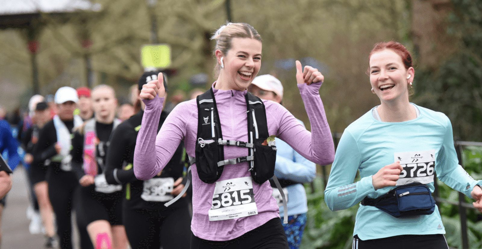 Two women smile and give thumbs up while running in a race, wearing numbered bibs and athletic clothing, with other runners visible in the background on a wooded path.