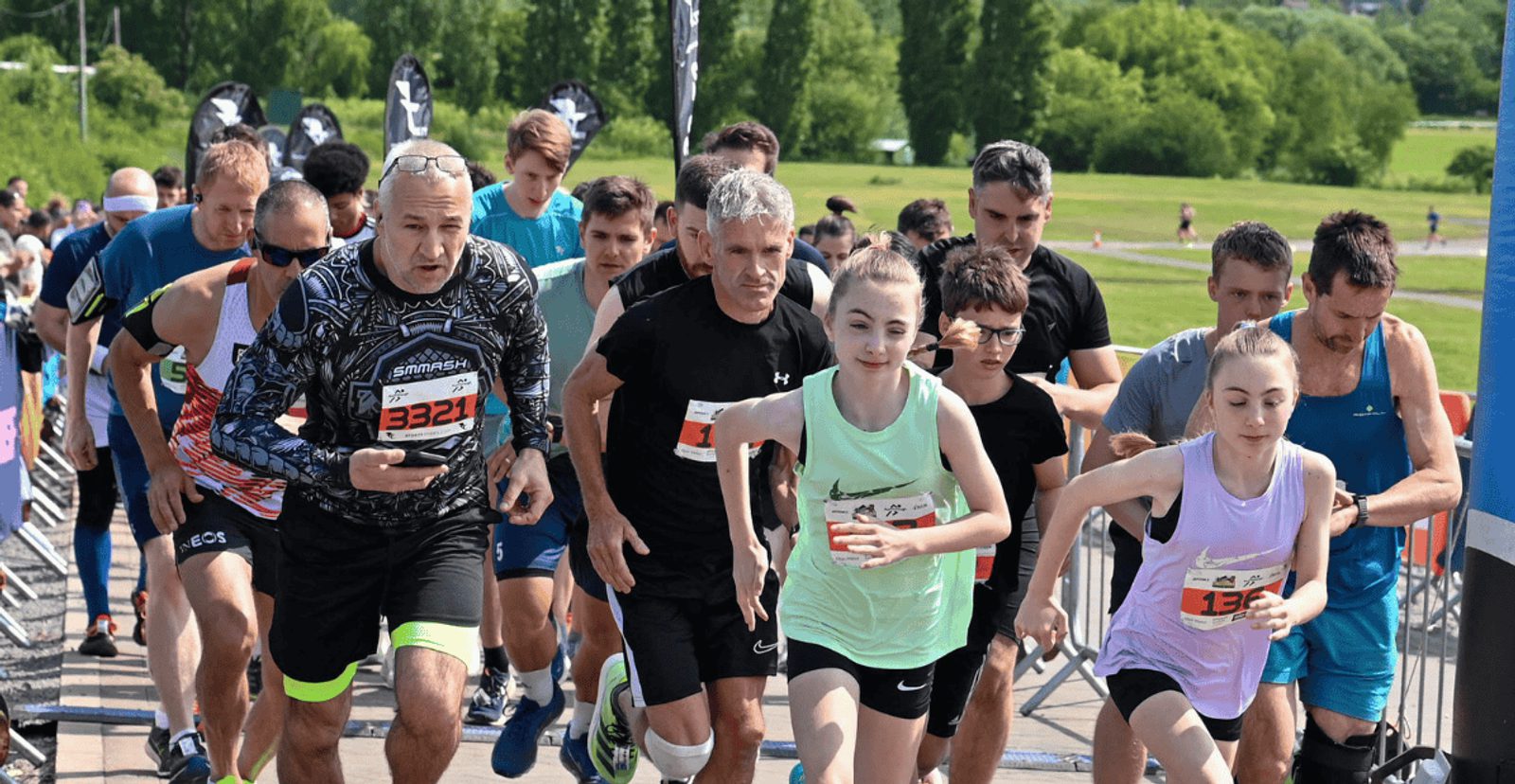 A group of runners, both men and women of various ages, start a race outdoors on a sunny day with green trees and grass in the background. Participants wear numbered bibs and athletic clothing.