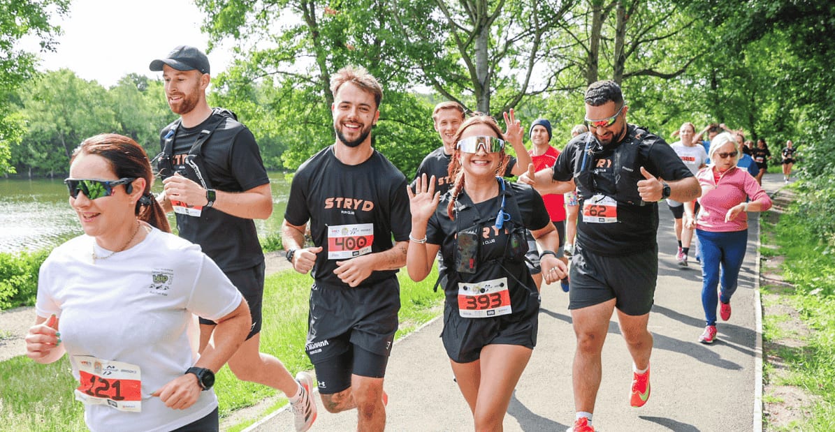 A group of people run together on a park path, smiling and waving. They wear athletic gear and race bibs, surrounded by green trees and a lake on a sunny day.
