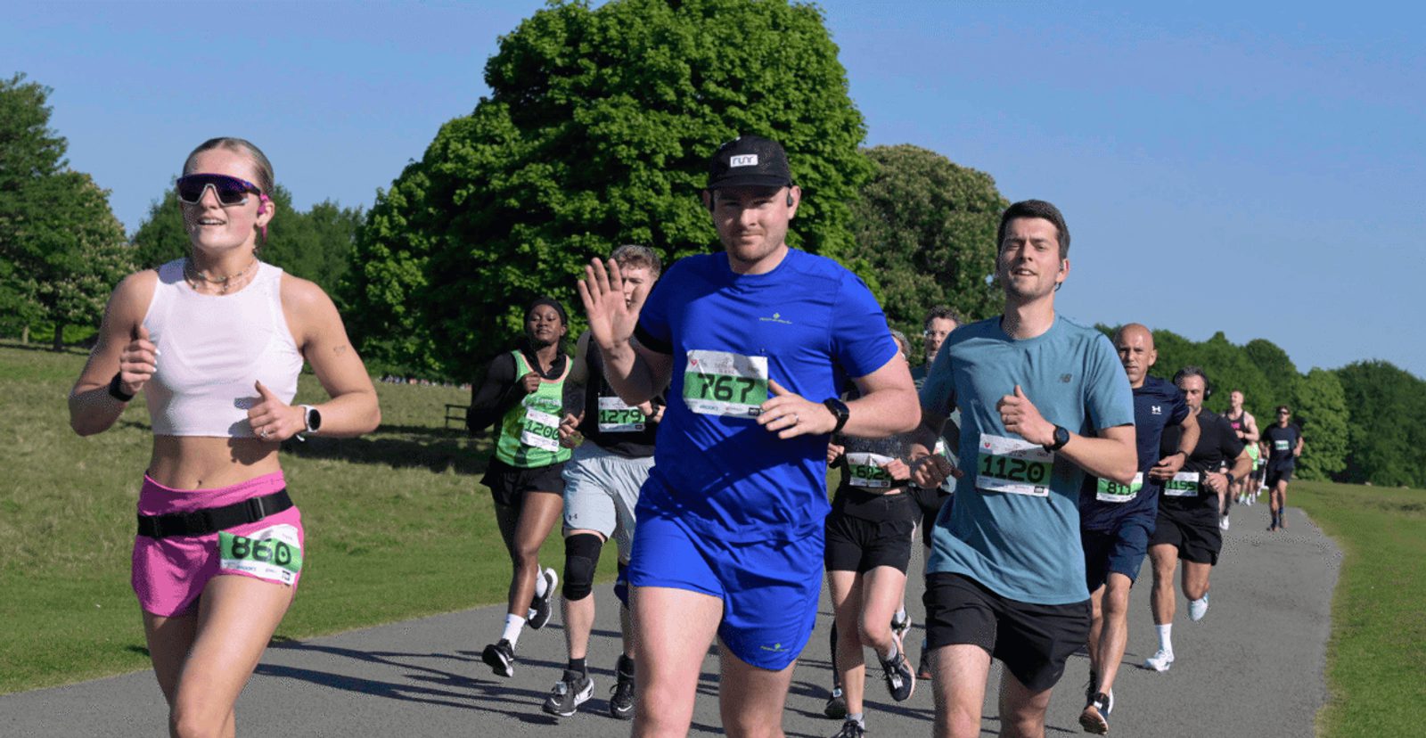 A group of people running in a race on a paved path in a park on a sunny day. The runners wear numbered bibs and athletic clothing, with trees and blue sky in the background.
