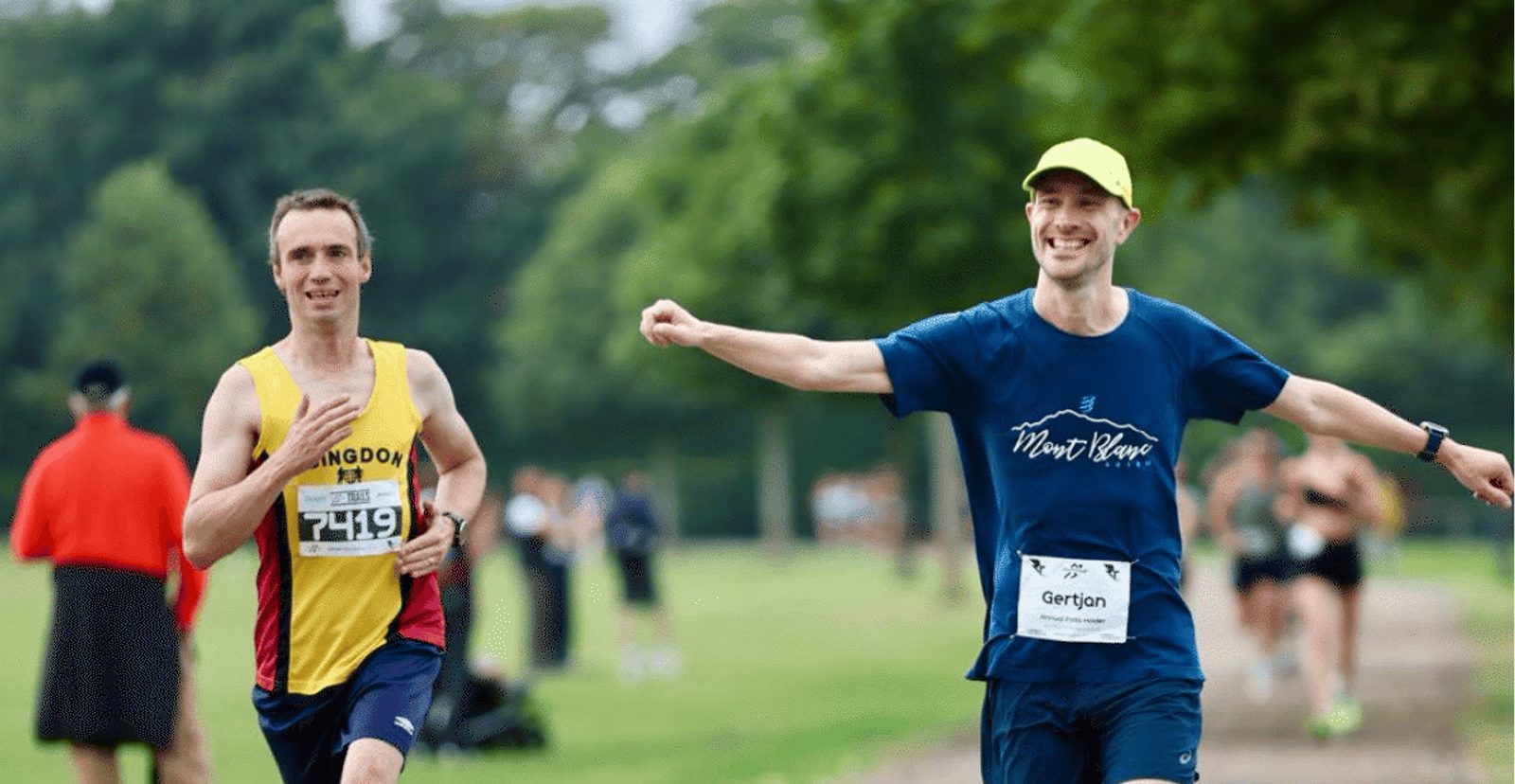 Two men are running outdoors in a race. One wears a yellow and red singlet with number 7419, and the other wears a blue "Mont Blanc" shirt and yellow cap, smiling and pointing. Other runners and trees are in the background.