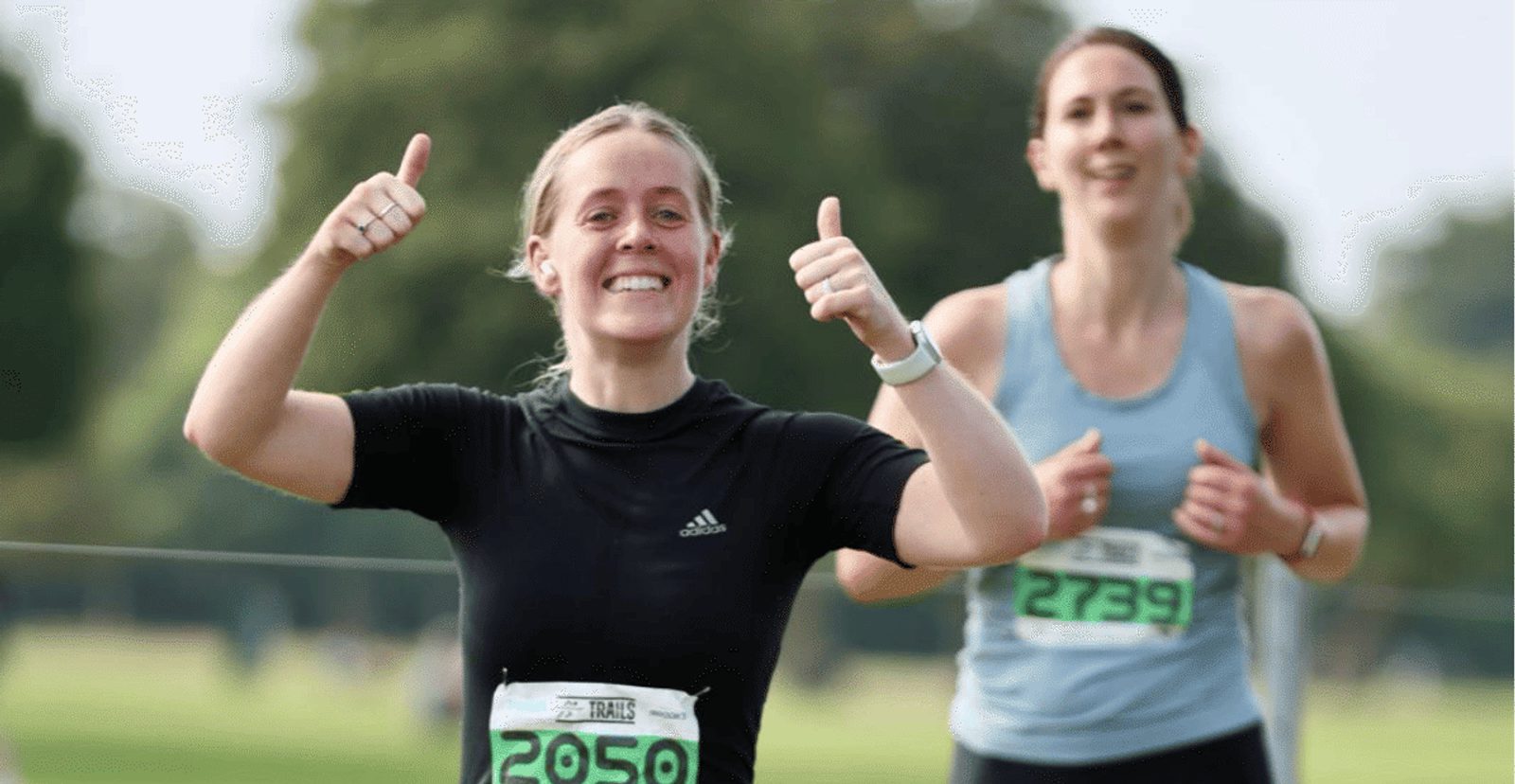Two women are running outdoors in a race. The woman in front, wearing a black shirt and race number 2059, is smiling and giving two thumbs up. The woman behind her wears a light blue tank top and race number 2739.