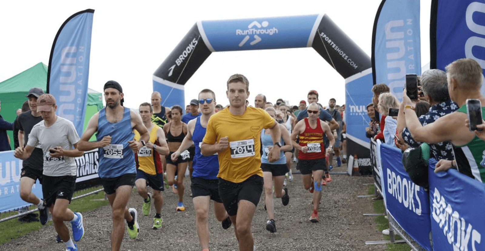 A group of runners, wearing numbered bibs, start a race under a black inflatable arch with "RunThrough" branding, surrounded by blue banners and cheering spectators on a gravel path.