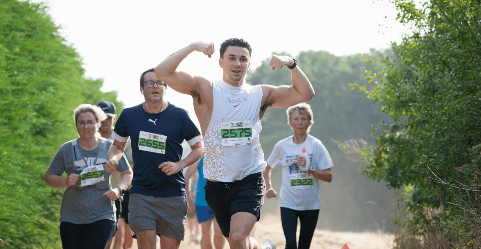 A group of runners participate in a race outdoors. The man in the front flexes his arms and smiles; others follow behind, wearing race bibs and athletic gear, running along a green, tree-lined path.