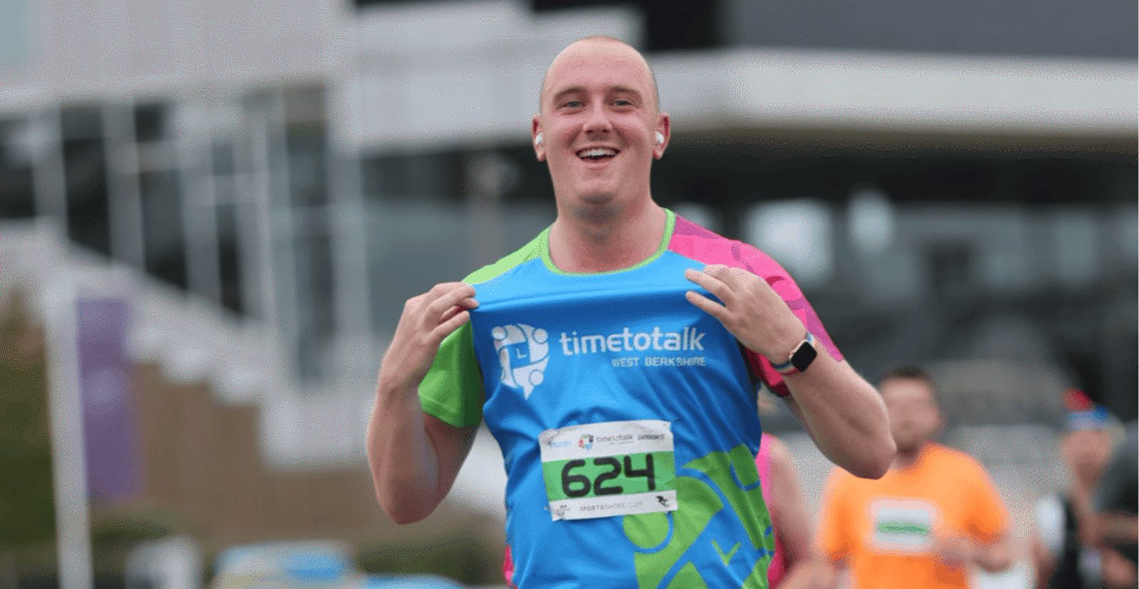 A smiling runner wearing a colorful "Time to Talk" shirt and bib number 624 jogs in a race, holding his shirt by the shoulders. Other runners and blurred buildings are visible in the background.