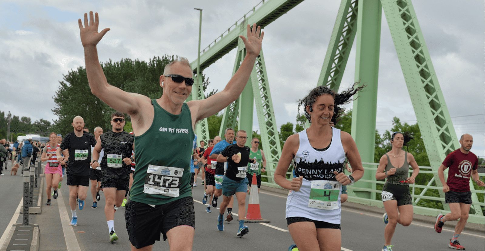 A group of runners cross a bridge during a race. A smiling man with sunglasses raises his arms high, while a woman next to him smiles and runs. Other participants and a green metal bridge structure are visible in the background.
