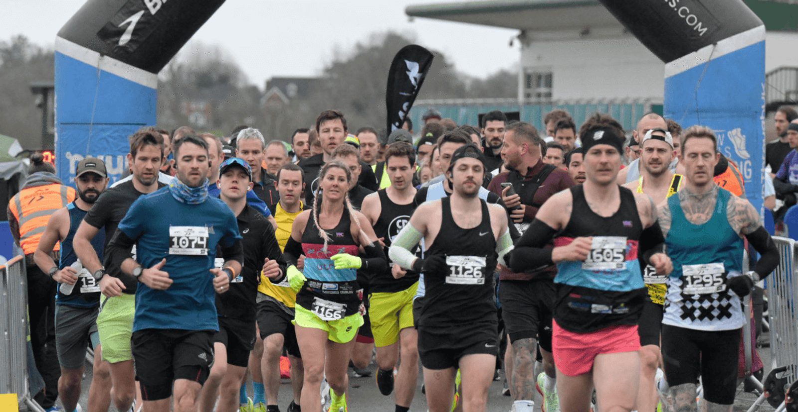 A large group of runners, wearing numbered bibs and athletic gear, start a race together under a blue inflatable arch on a cloudy day. Spectators and event staff stand behind barriers along the route.