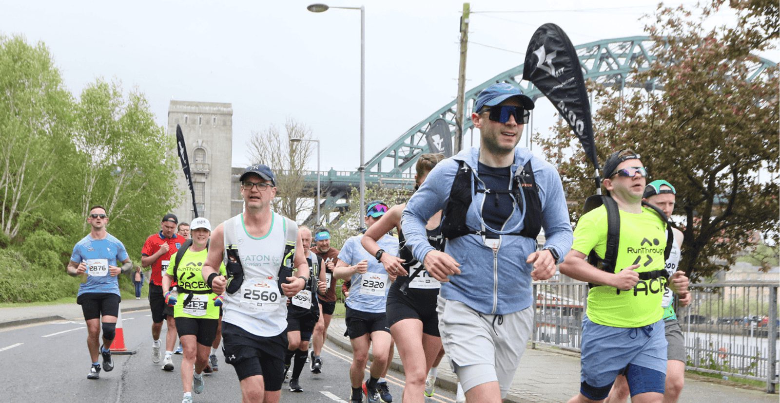 A group of marathon runners on a city street, wearing race bibs and athletic gear. A bridge and trees are visible in the background, and the weather appears cool and overcast.