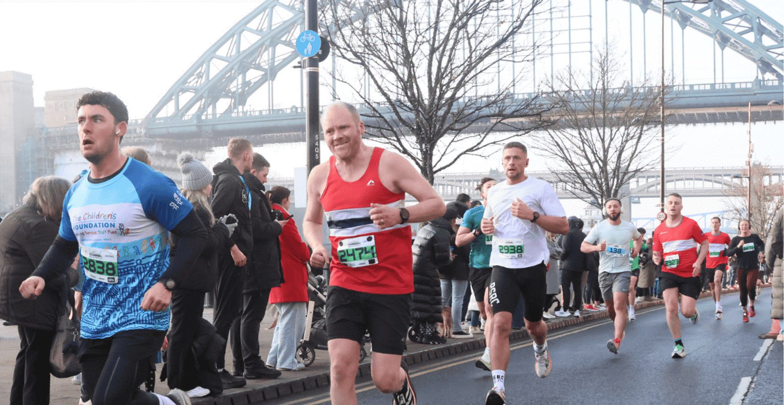Runners participate in a road race near a bridge, with spectators lining the street. Some runners wear numbered bibs and athletic gear, while the background features a large arched bridge and bare trees.