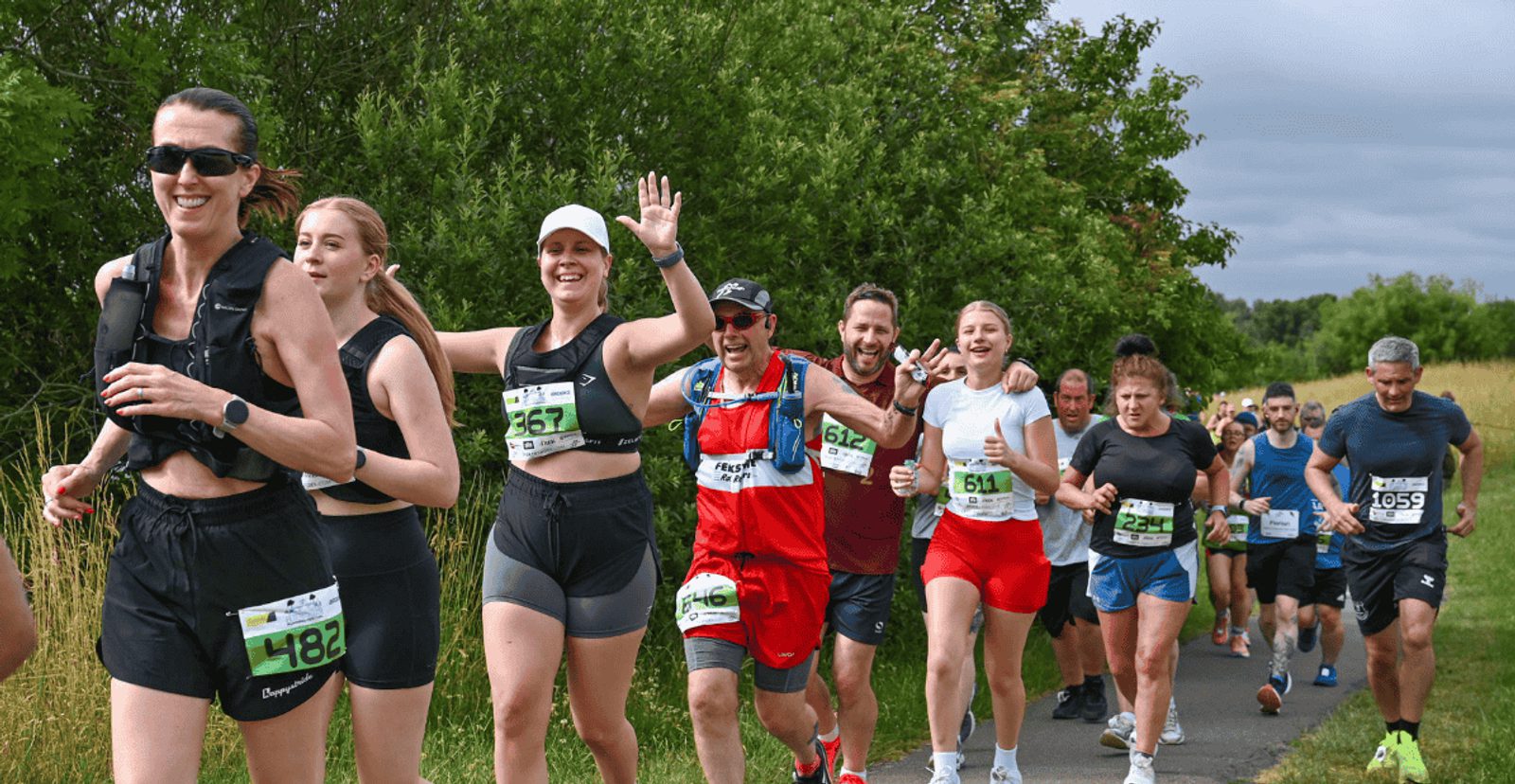 A group of smiling runners, wearing race bibs and athletic gear, jog together along a paved path bordered by grass and trees during a race or fun run on a partly cloudy day.