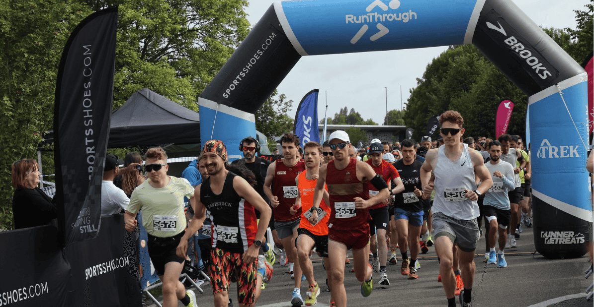 A group of runners starts a race, passing under a blue inflatable archway marked "RunThrough" and sponsor logos, with spectators watching from the sidelines. The runners wear athletic gear and numbered bibs. Trees line the background.