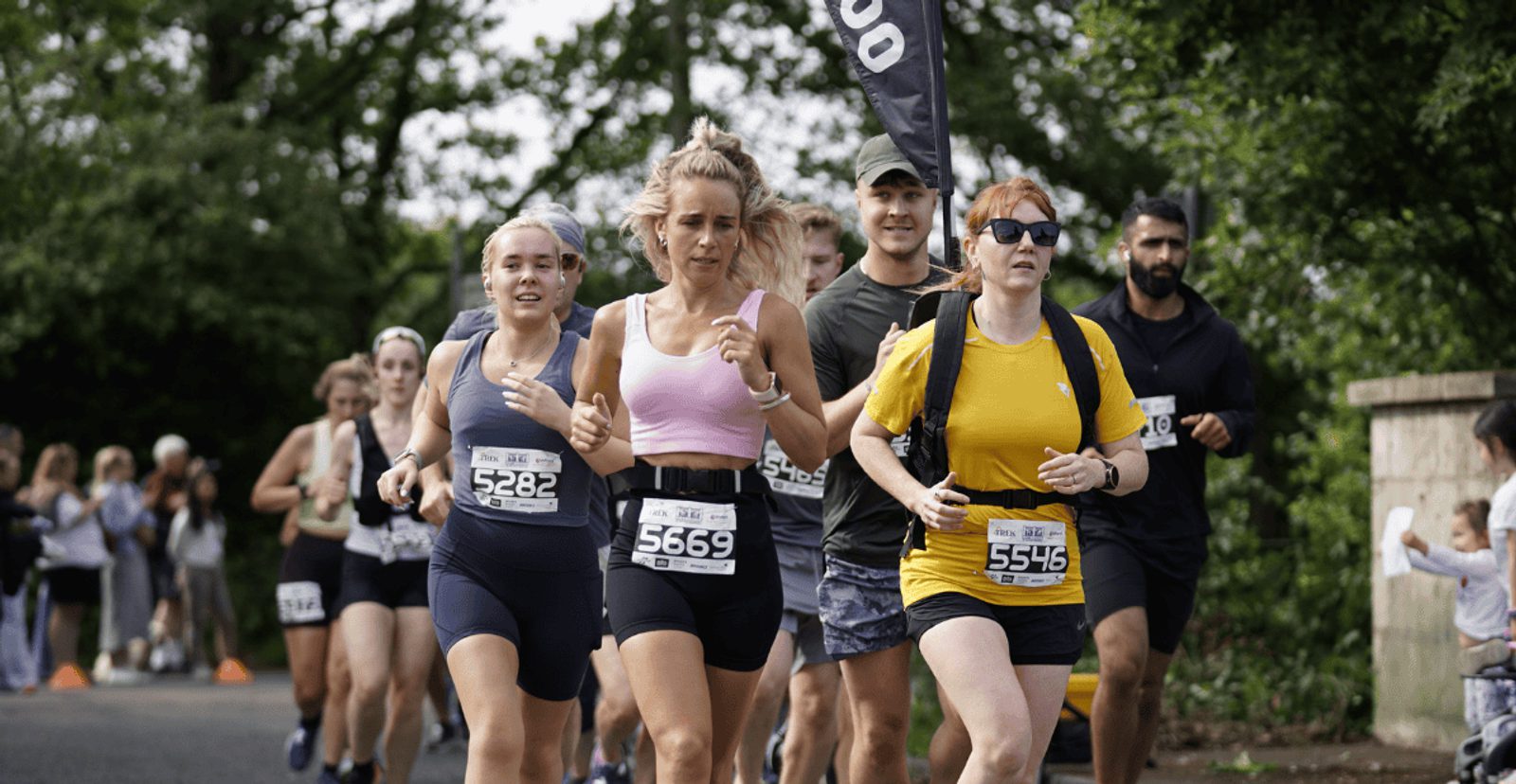 A group of runners with race bibs jogs outdoors in a marathon event, surrounded by greenery and spectators cheering from the side. The lead runners include women in athletic gear.