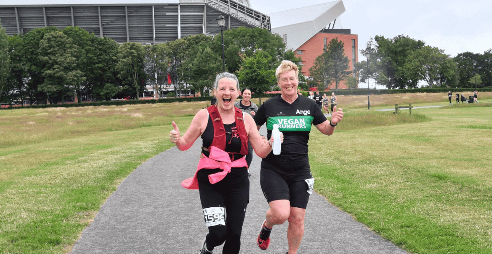 Two smiling runners, one wearing a pink jacket and race bib, the other in a black "Vegan Runners" shirt, give thumbs up while running on a park path near a large stadium on a cloudy day.