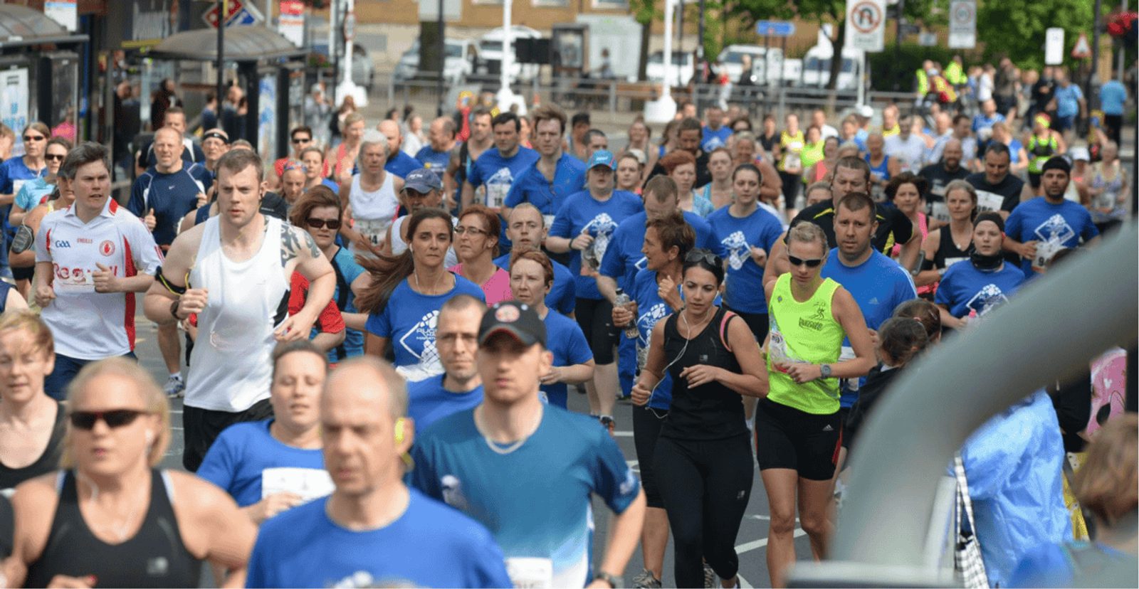 A large group of runners participates in a road race on a city street. Most are wearing blue or light athletic clothing, and spectators can be seen in the background. The event takes place in daylight.