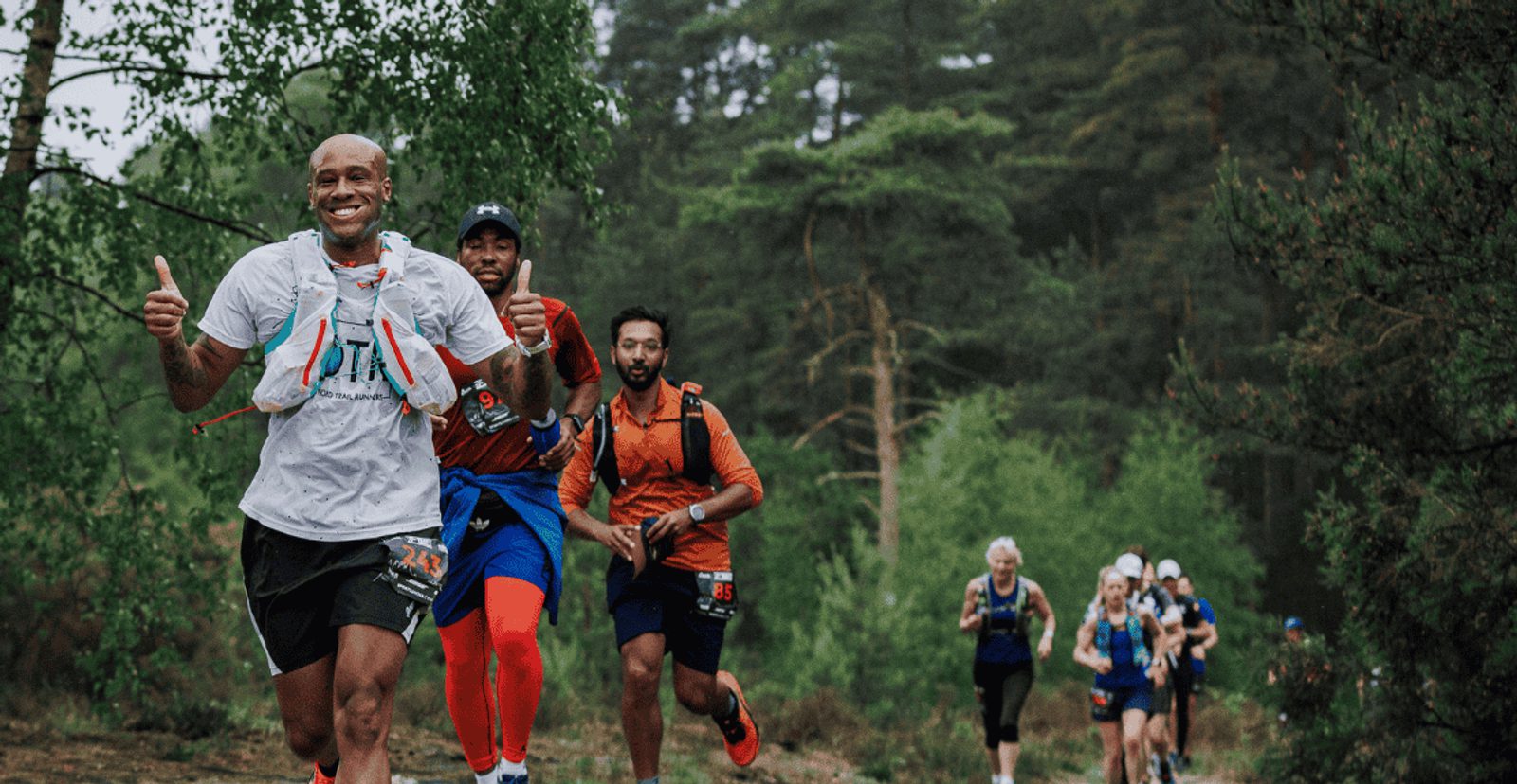 A group of trail runners, led by a smiling man giving two thumbs up, run through a forested path. The runners behind him wear athletic gear and hydration packs, surrounded by lush green trees.