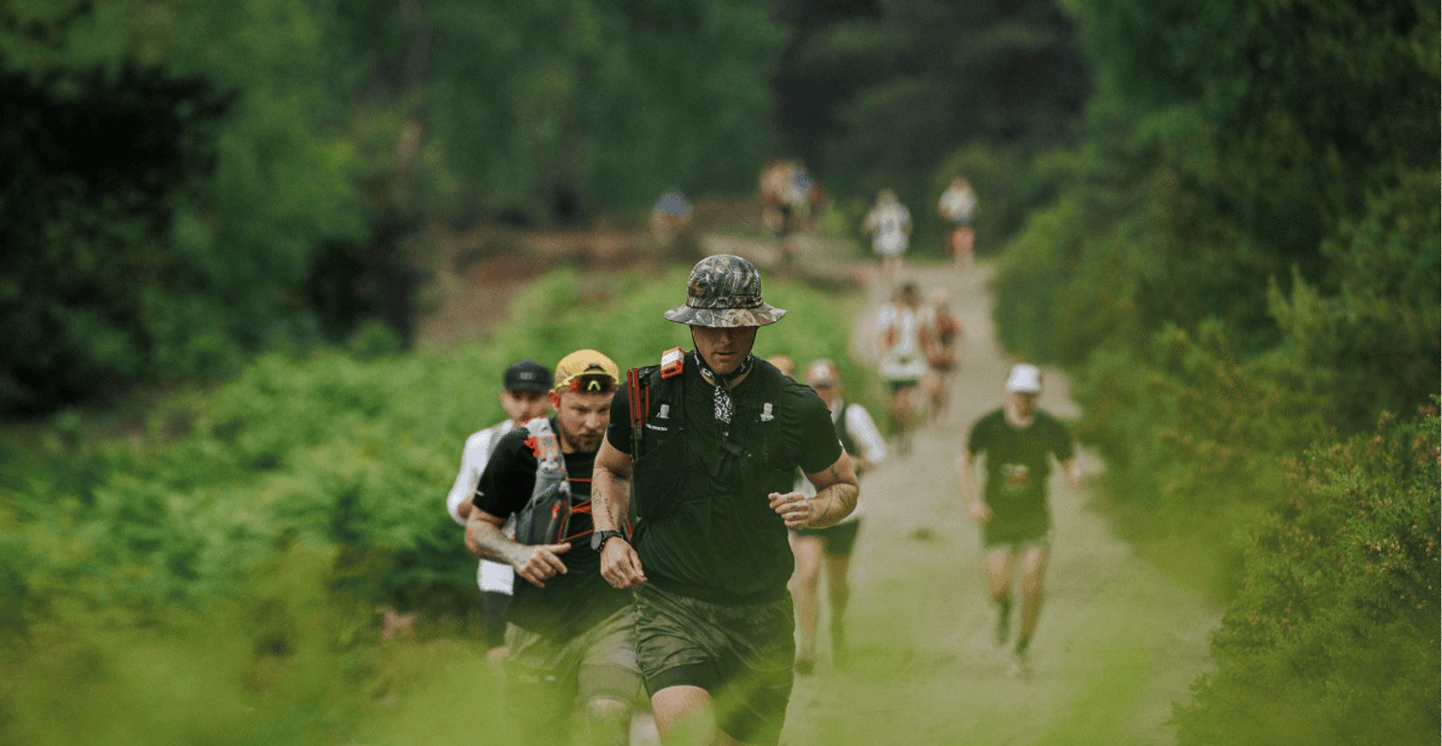 A group of trail runners in athletic gear jog through a lush, green forest path, with the lead runner wearing a camouflage hat and the rest following closely behind.