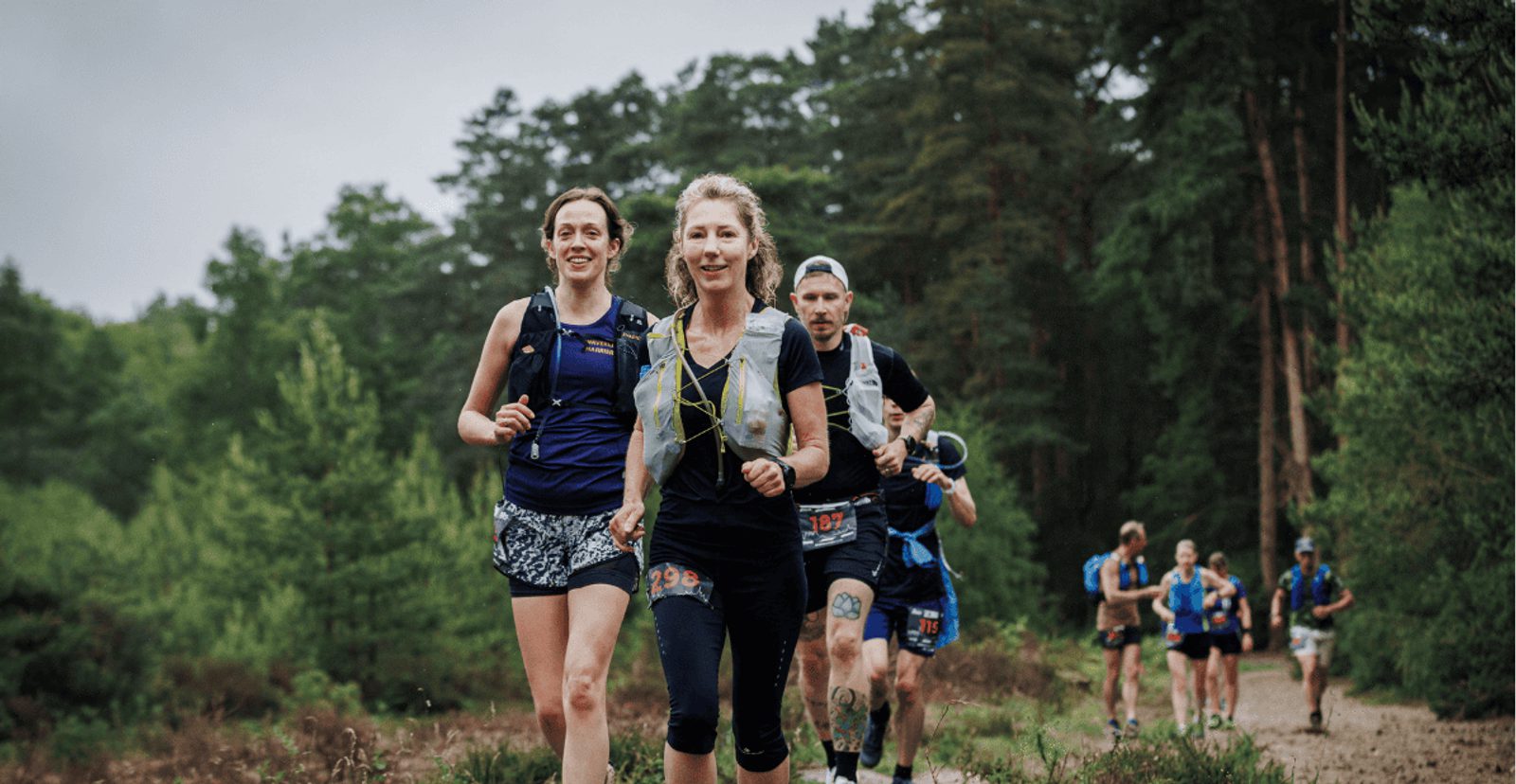 A group of trail runners, including two women in front, jog along a forest path surrounded by green trees, wearing athletic gear and numbered race bibs. The weather appears overcast.