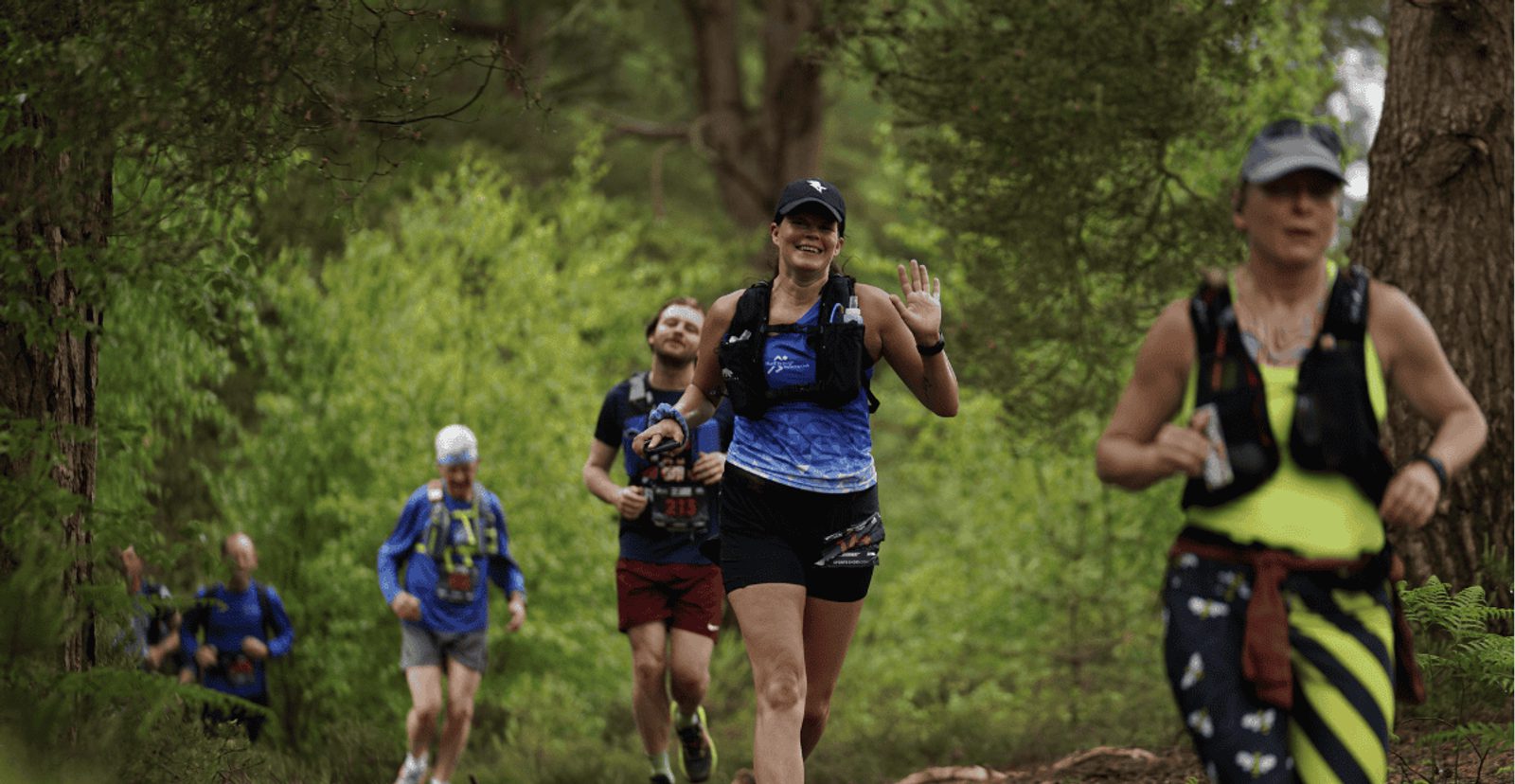 Four people run on a forest trail surrounded by green trees. The woman in front, smiling and waving, wears a blue tank top and cap. Other runners follow behind, wearing athletic gear and hydration packs.