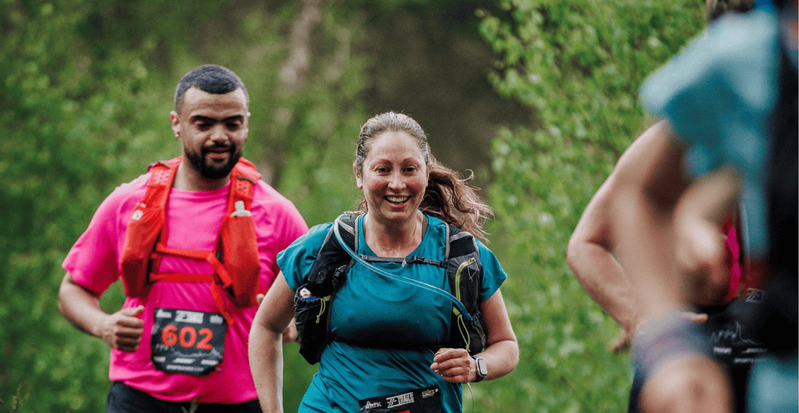 A woman and man in athletic gear are running outdoors on a trail, smiling. The woman is in front wearing a teal shirt and black vest; the man behind her wears a pink shirt and red hydration pack. Greenery is in the background.
