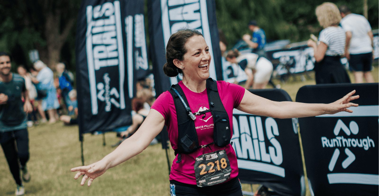 A smiling woman in a pink shirt and race bib 2218 stretches out her arms as she finishes a trail run, with banners and other participants in the background.
