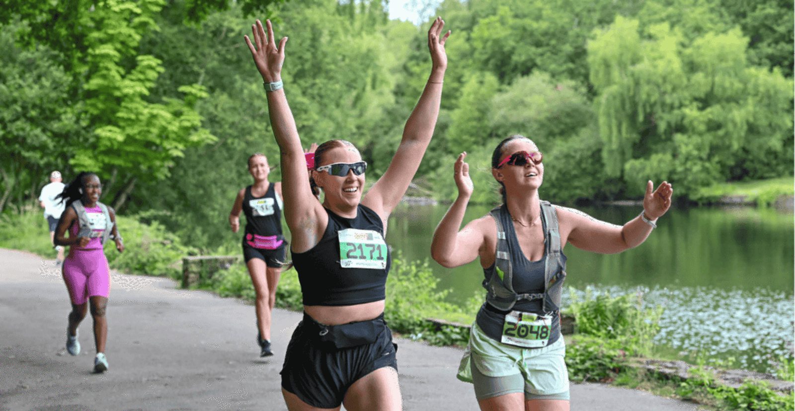 Two women with race bibs smile and raise their arms in celebration as they run on a path by a lake, with two other runners behind them, surrounded by trees and greenery.