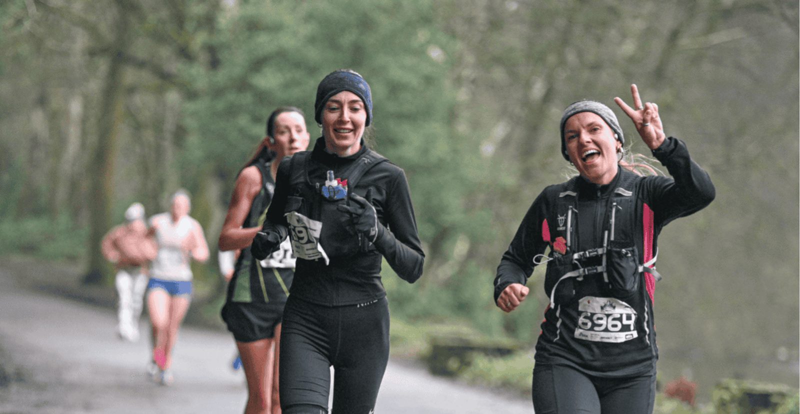 Two women in running gear, smiling and jogging on a forest trail during a race. One holds up a peace sign. Other runners are visible in the background on the wet path.