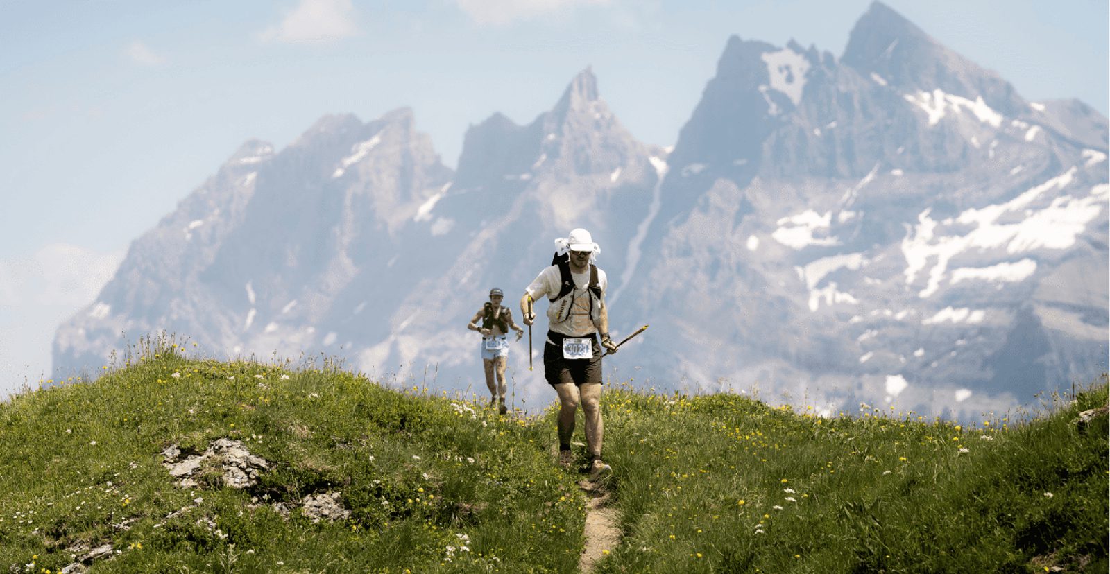 Two hikers walk along a narrow grassy trail with wildflowers, surrounded by green hills and tall, snow-patched mountains in the background under a partly cloudy sky.