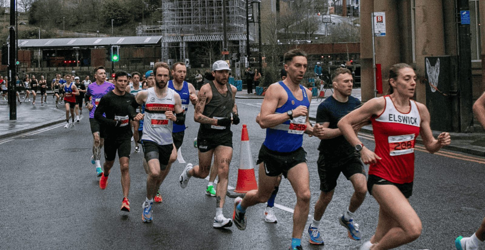 A group of runners, wearing race bibs and athletic gear, participates in a road race on a wet city street. Some wear club jerseys; orange traffic cones line the course and spectators are visible in the background.