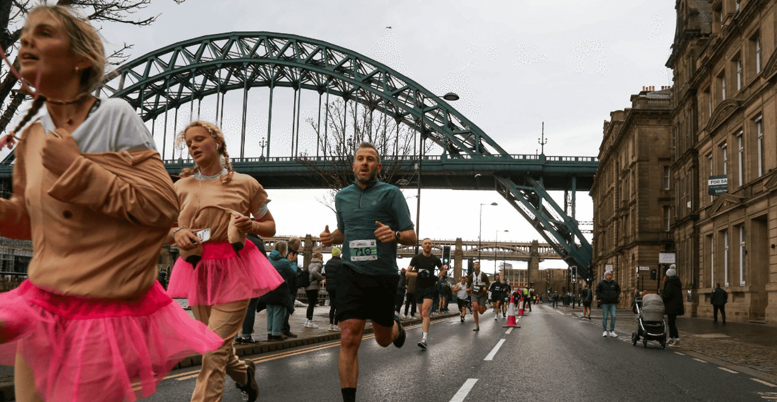 Runners, including people in pink tutus, participate in a road race on a city street with a large arch bridge in the background. Spectators watch from the sidewalk under an overcast sky.