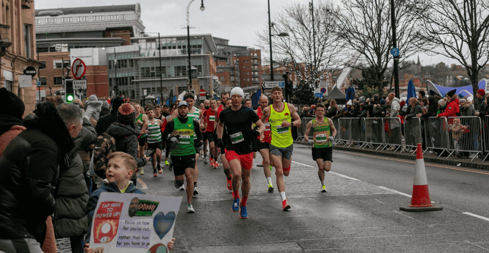 A group of marathon runners in colorful athletic wear race on a city street lined with cheering spectators behind barriers on a cloudy day. A child holds a motivational sign in the foreground.