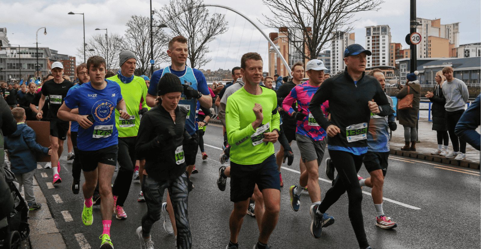 A group of runners in athletic gear participate in a race on a city street, with buildings, trees, and spectators visible in the background. Some runners wear bright neon shirts and race bibs.