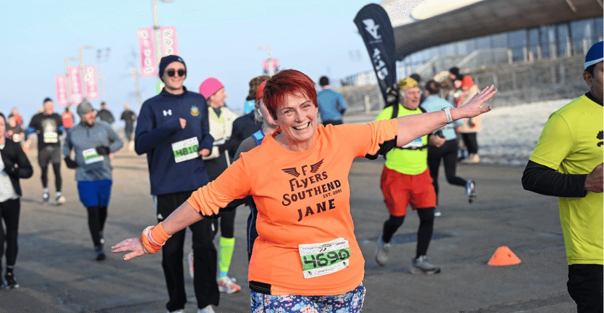 A woman with short red hair, wearing an orange "Flyers Southend Jane" shirt, smiles with arms outstretched while running in a road race among other runners on a cool day.