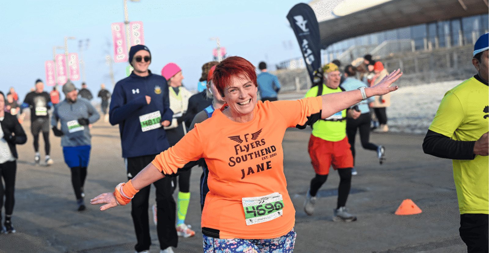 A woman with short red hair, wearing an orange "Flyers Southend Jane" shirt, smiles with arms outstretched while running in a road race among other runners on a cool day.