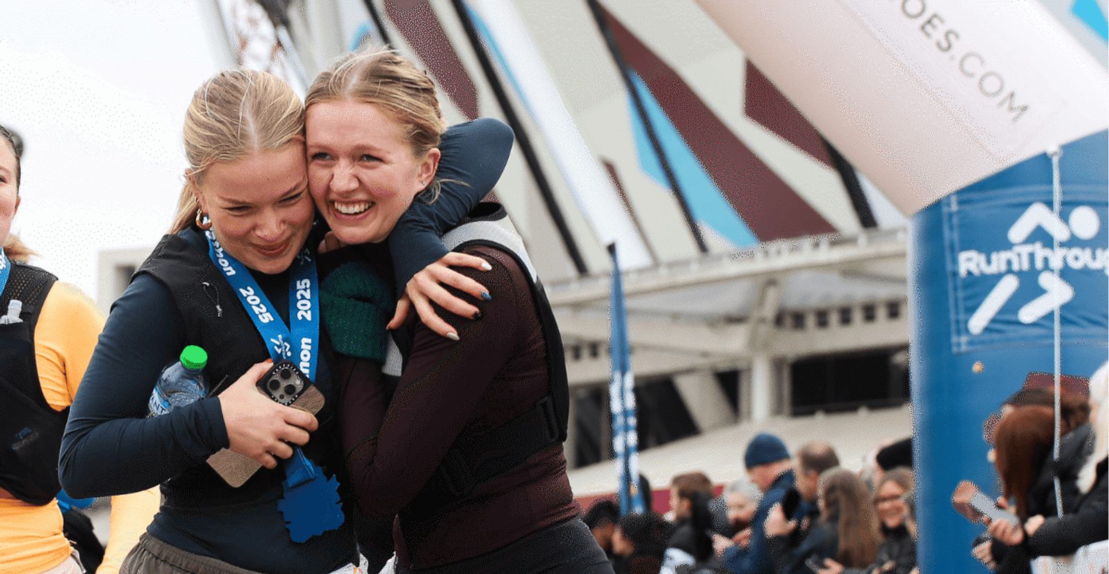 Two women smiling and hugging at the finish line of a race event, one wearing a medal around her neck and holding a water bottle, surrounded by other participants and onlookers.