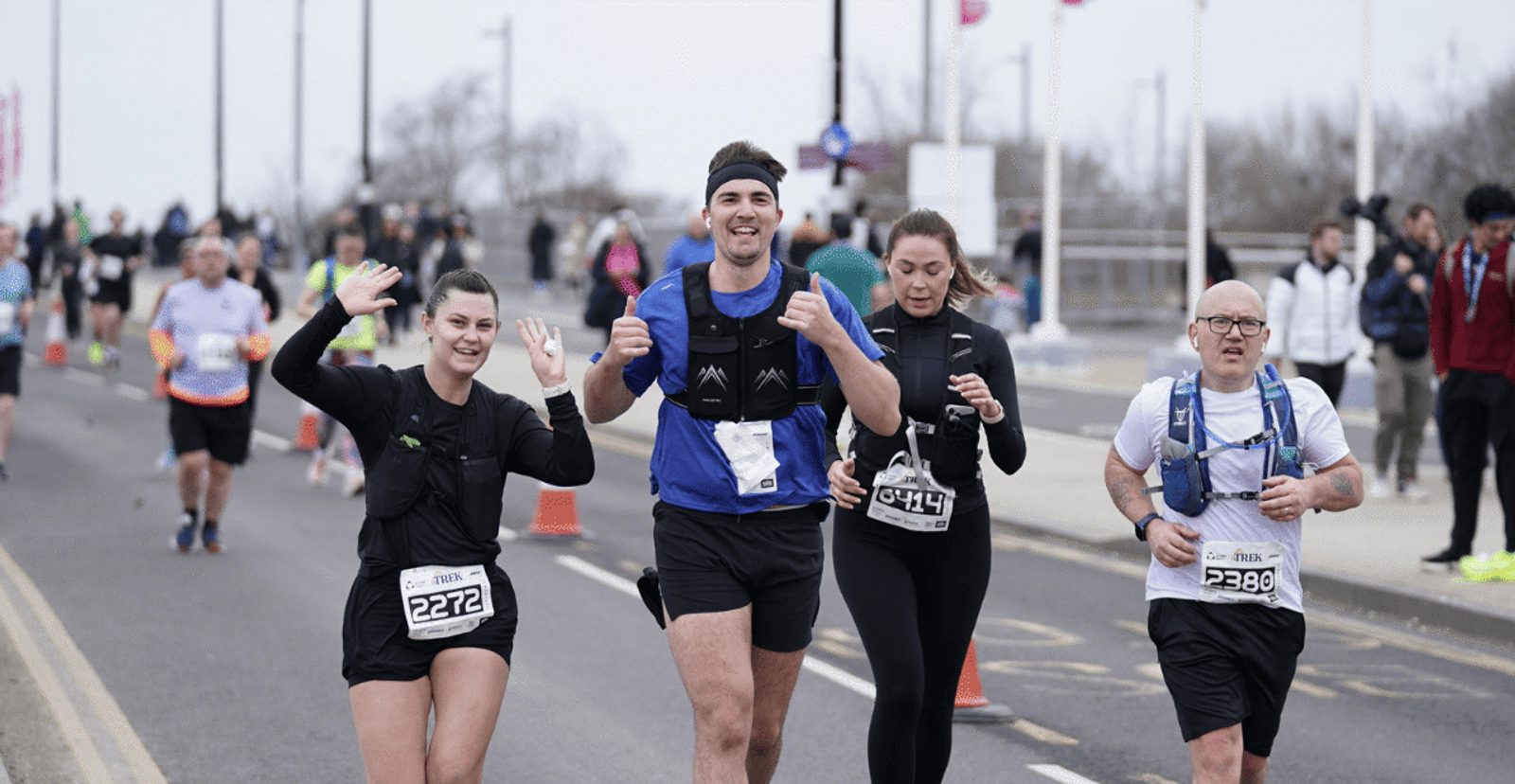 A group of runners smile and gesture to the camera while participating in a road race. Other runners and spectators are visible in the background on a cloudy day.