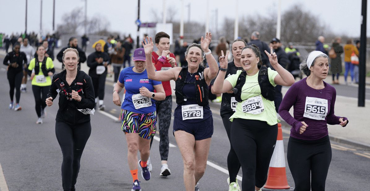A group of runners participate in a road race. Some are smiling and waving at the camera, while others focus on running. The weather appears cool and overcast, and more runners are visible in the background.