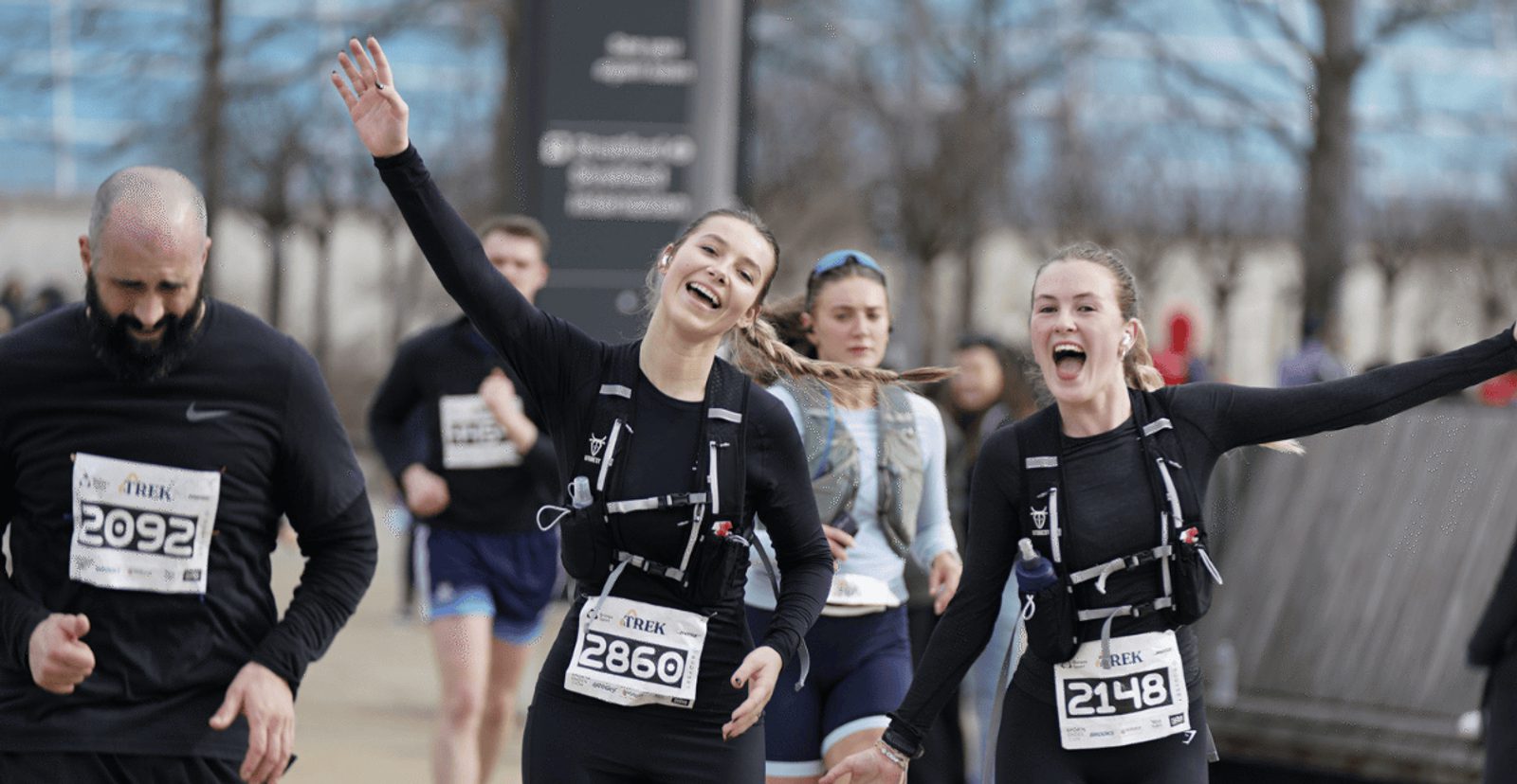 A group of runners wearing race bibs smile and cheer as they participate in a marathon. Two women in front raise their arms in excitement while a man runs beside them. Other runners are visible in the background.